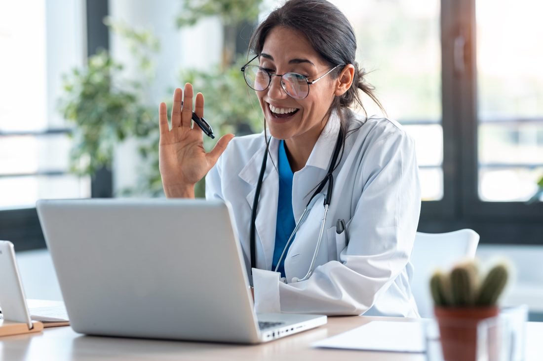 Smiling doctor in a white coat waving during an online video consultation on a laptop